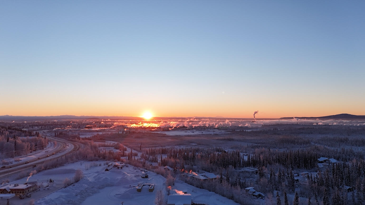 Low sun over a snowy developed valley with roads, buildings, trees, and rising plumes.