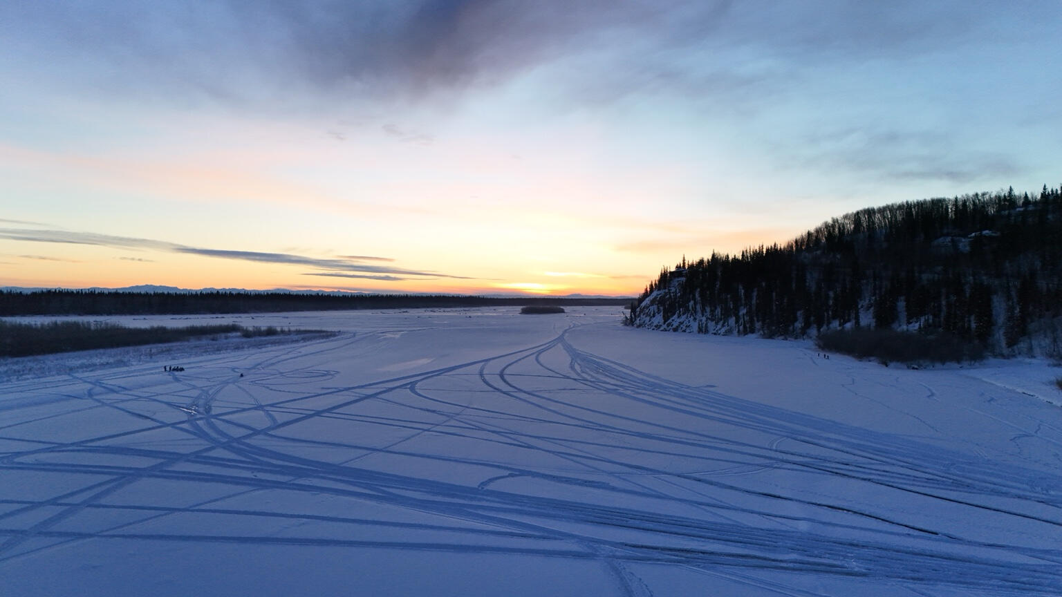 Tracks across a snow-covered open landscape beside a wooded bluff at sunset.