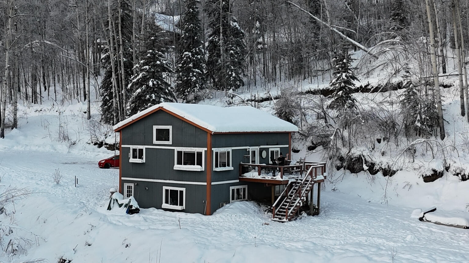Snow-covered two-story house with a deck and stairs in a wooded setting.